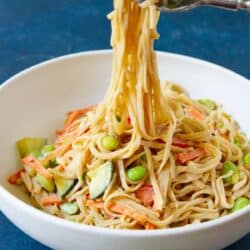 A fork lifting rice noodles out of white bowl filled with peanut noodles and vegetables.