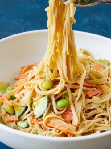 A fork lifting rice noodles out of white bowl filled with peanut noodles and vegetables.