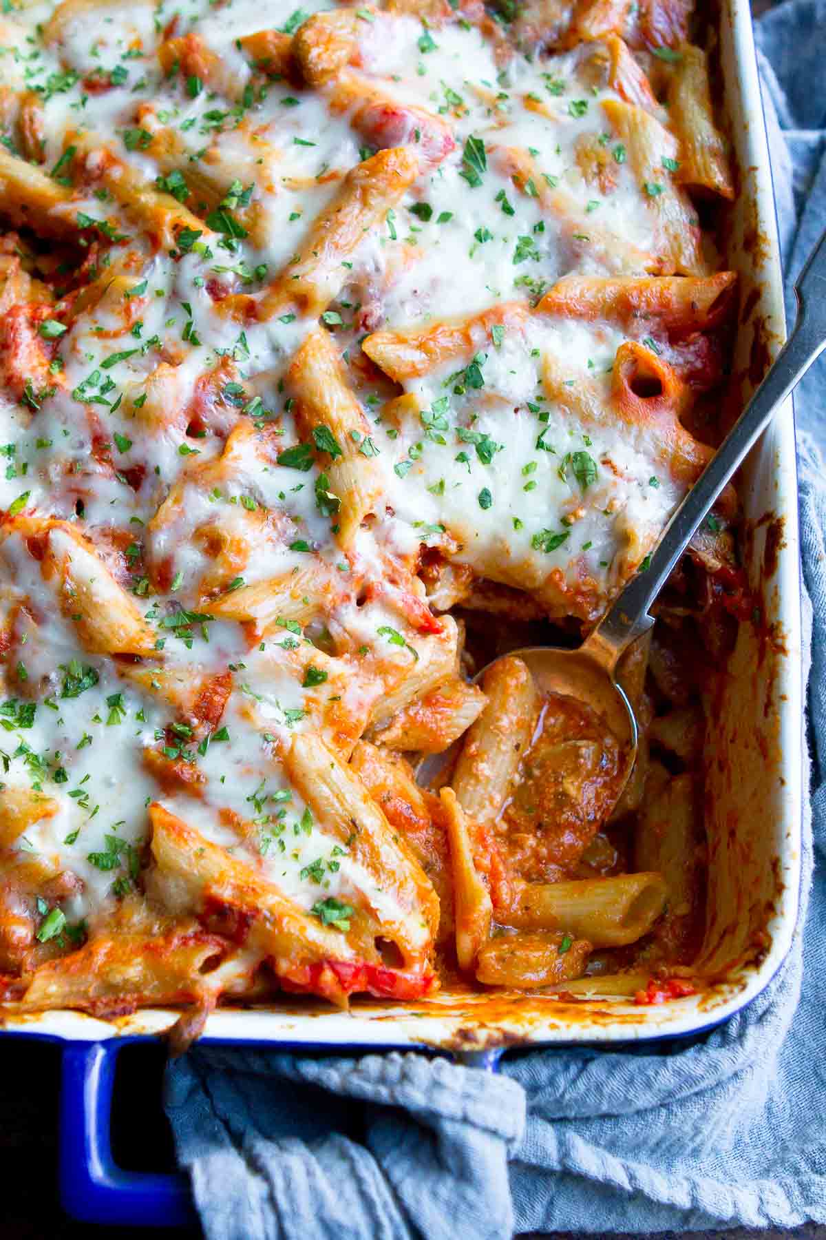 A serving spoon dipping into a large casserole dish filled with baked ziti topped with melted cheese.