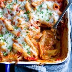 A serving spoon dipping into a large casserole dish filled with baked ziti topped with melted cheese.