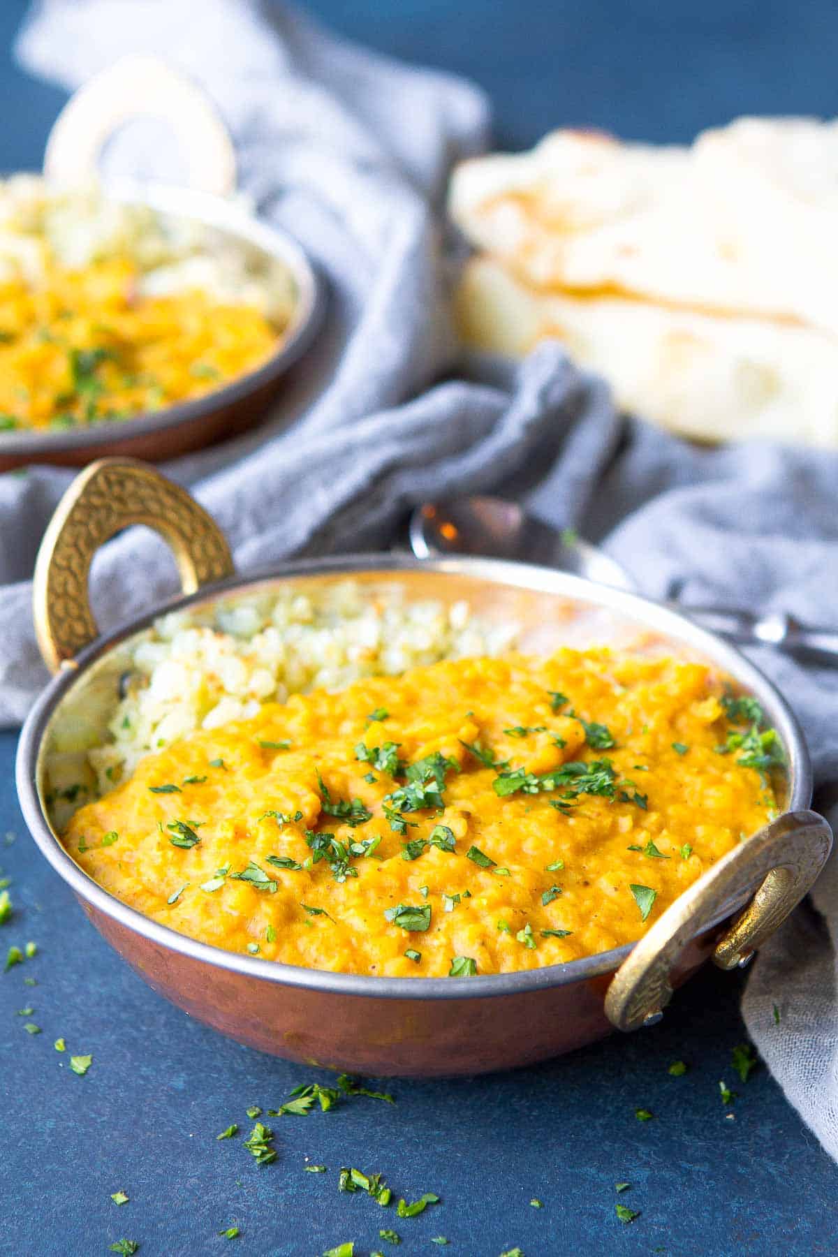 Two brass bowls with handles, fill with red lentil dal and rice. Resting on a gray napkin.