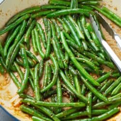 Glazed green beans in a large ceramic skillet, with tongs.