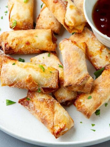 Cooked mini egg rolls and a bowl of pepper jelly on a white plate.