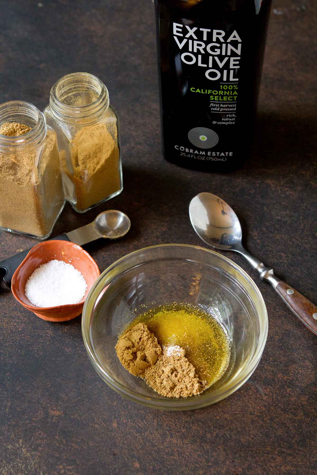 Spice rub in a glass bowl, surrounded by spice bottles and olive oil.