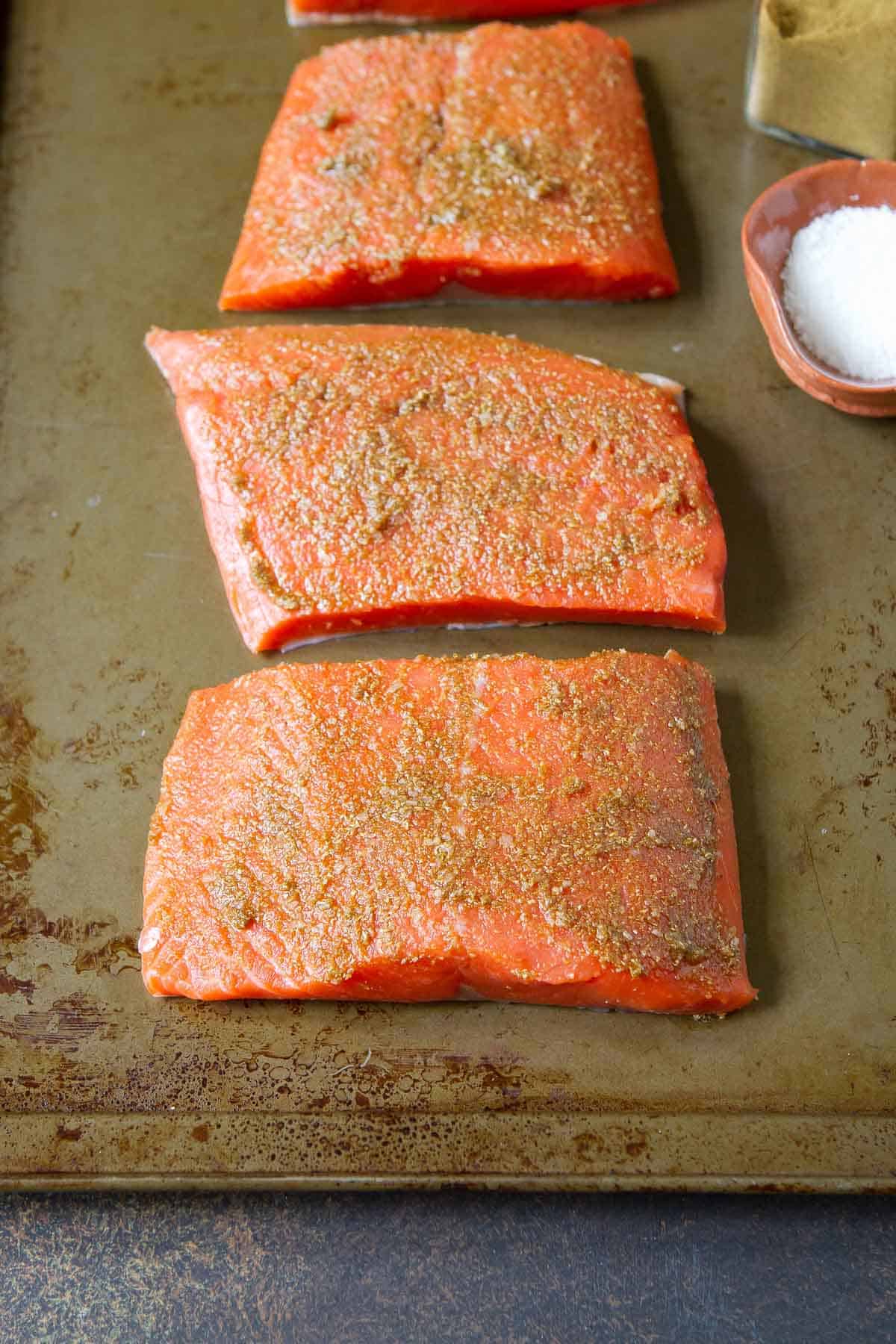 Raw salmon fillets with spice rub, lined up on a baking sheet.