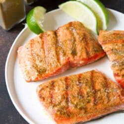 Three grilled salmon fillets on a white plate with lime wedges.