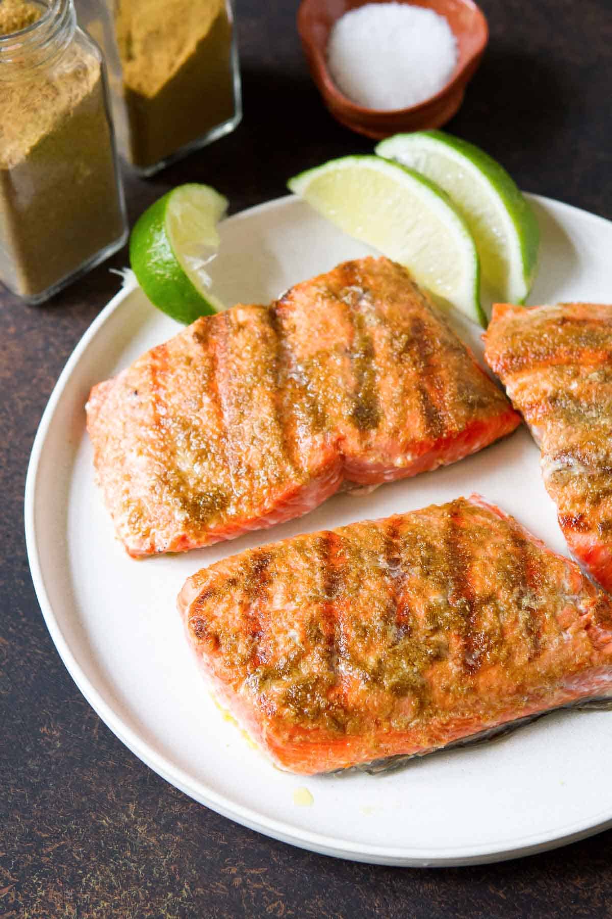 Three grilled salmon fillets on a white plate with lime wedges.