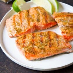 Three grilled salmon fillets on a white plate with lime wedges.