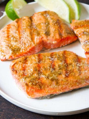 Three grilled salmon fillets on a white plate with lime wedges.