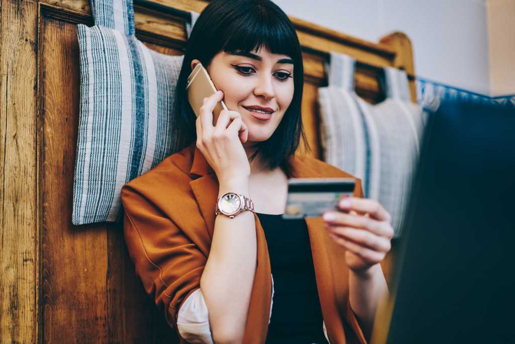 Positive woman looking at number on credit card and confirm purchase via telephone call to customer service, smiling hipster girl making payment via smartphone conversation while resting indoors