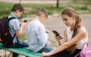 young girl and two boys sitting on a bench lwith smartphones in their hands