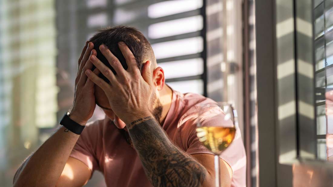 Junger fitter Mann hält sich den Kopf, daneben steht ein Glas Wein.