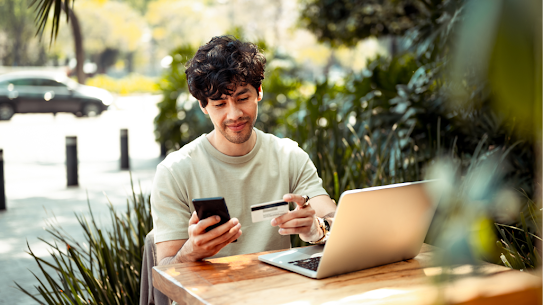 A photo of a man using his phone, laptop and a credit card.