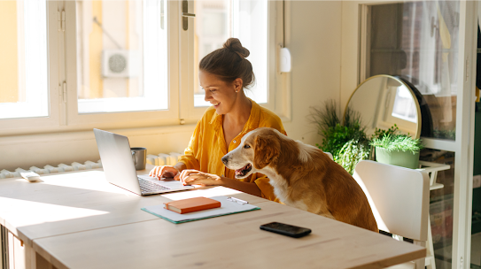 A photo of a woman working at home on her laptop with her dog next to her