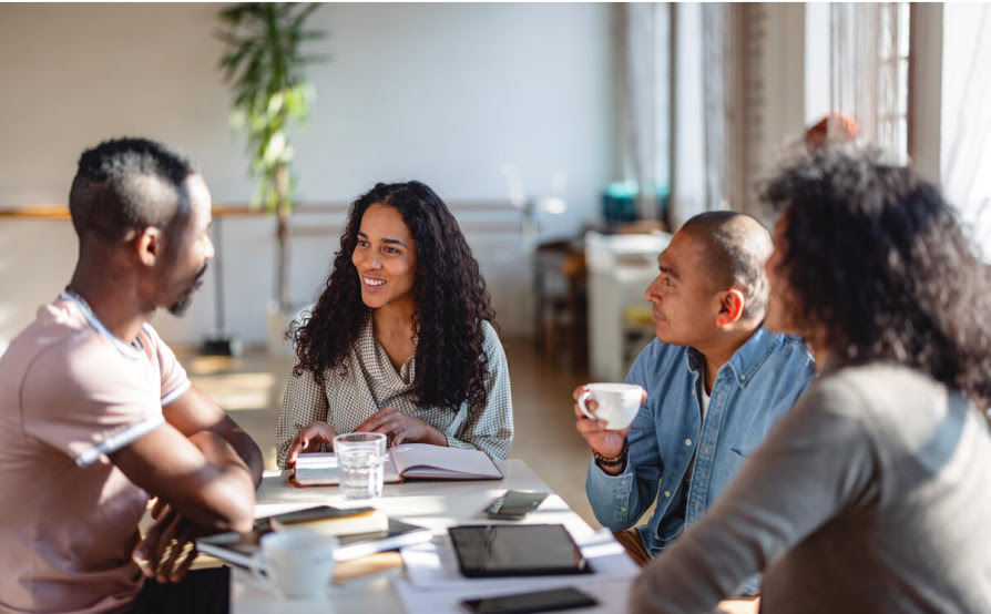 Four team members sit at a table having a coffee-work meeting with notebooks, tablets, and phones across the table.