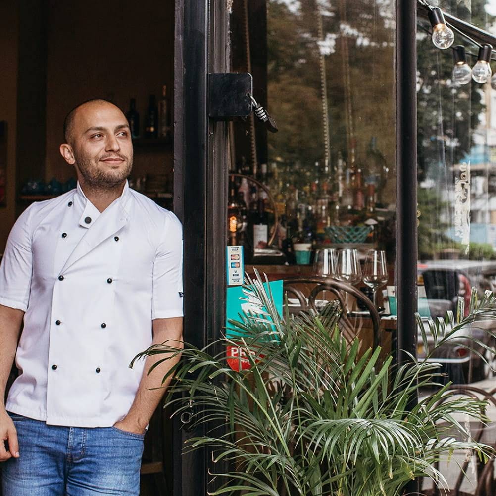 Man with chef clothes leaning in the doorway of a restaurant.