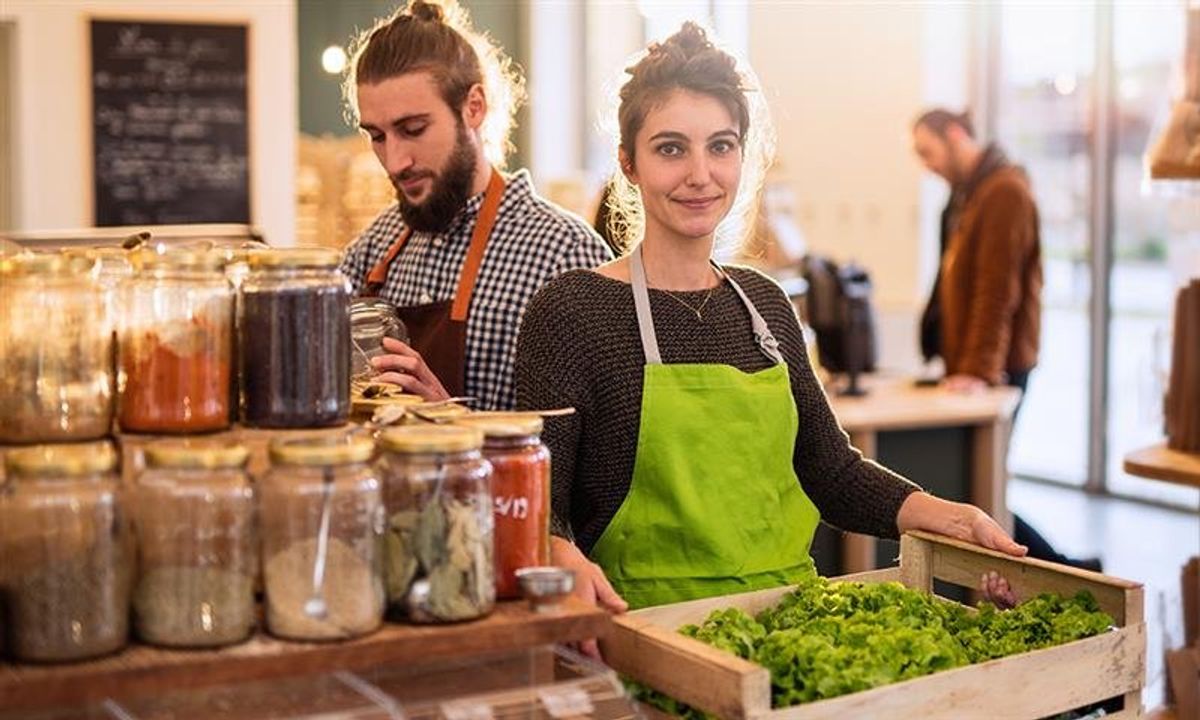 A woman holding a crate of leafy greens and a man behind her taking out spices from a jar