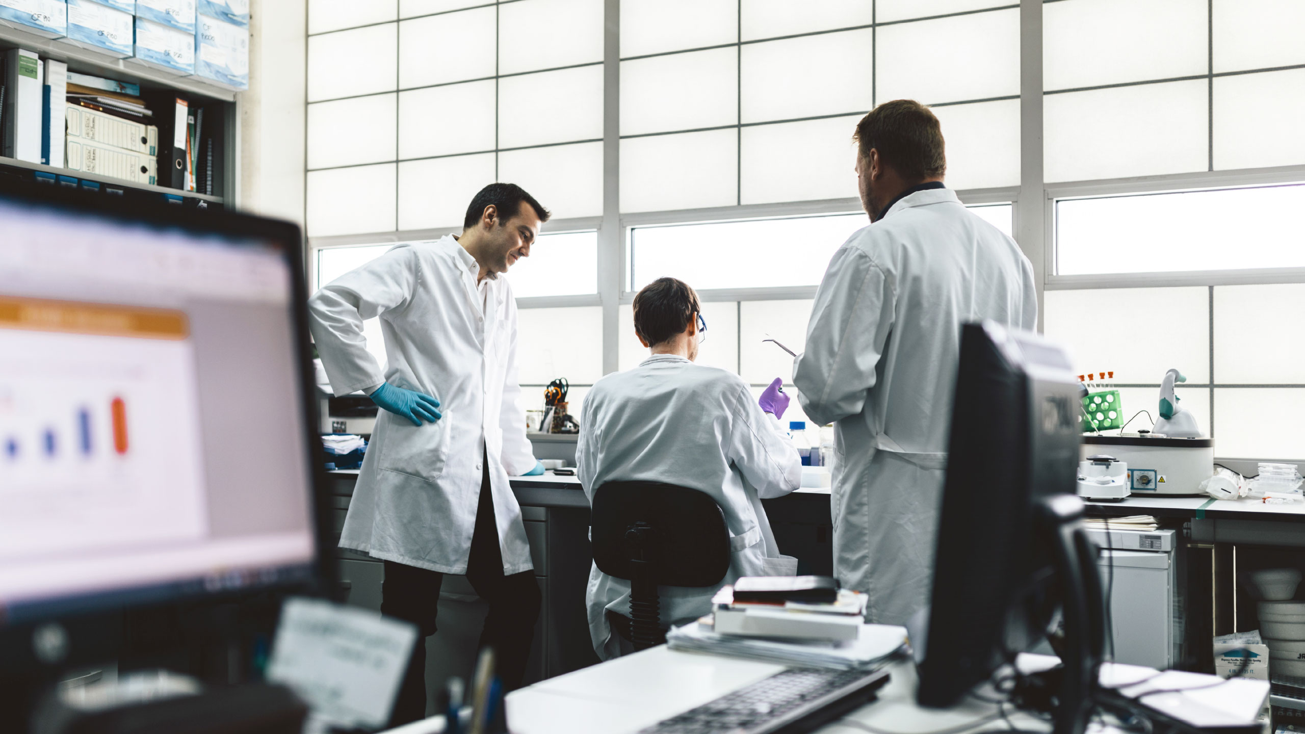 Scientist is seated with back facing camera with two scientists standing on either side