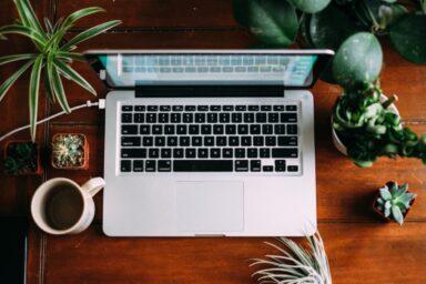 An open laptop sits on a desk alongside potted plants and a cup of coffee, illustrating the convenience and accessibility of online therapy.