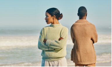 A man and woman stand on a beach, arms crossed, backs to each other in passive-aggressive manner.