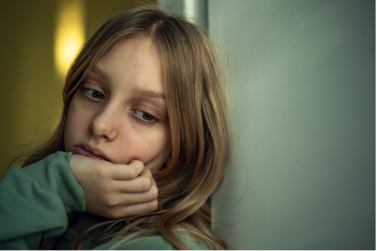 A young girl leans against a wall, sad eyes looking down, her expression reflecting a sense of anhedonia or loss of pleasure