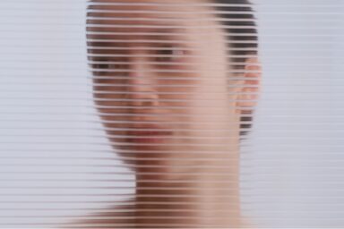 A woman gazes thoughtfully through window blinds, with a softly blurred background enhancing the sense of cognitive dissonance