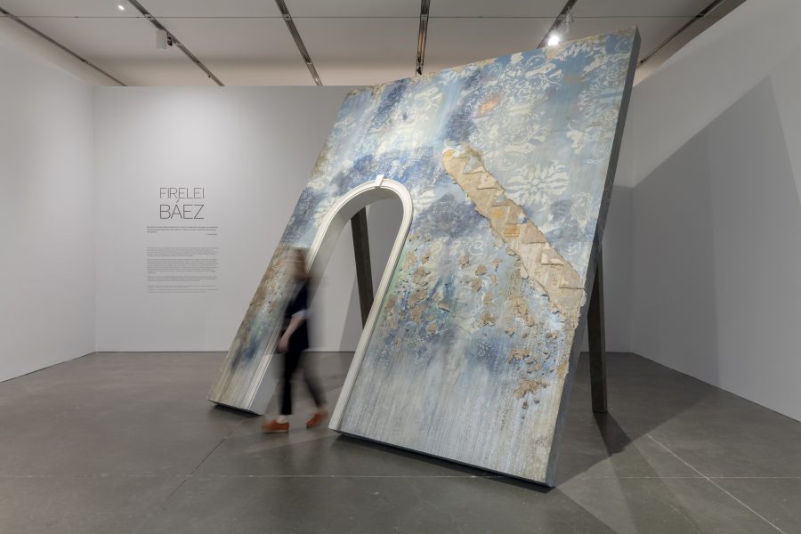 A blurry figure walks out of a blue sculptural arch in a gallery