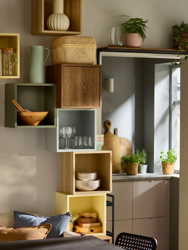 A vertical wall storage with open and closed EKET cabinets in various colours, filled with glasses and FÄRGKLAR plates.