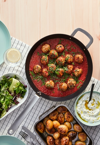 A pan with mince balls, made from HUVUDROLL Plant balls, in tomato sauce and an oven tray with roasted potatoes.