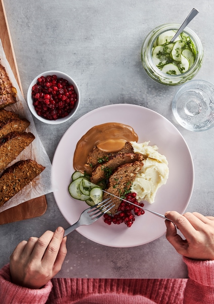 A knife and fork about to dig into a plate of meatloaf, sliced cucumbers, lingonberries, cream sauce and mashed potatoes.