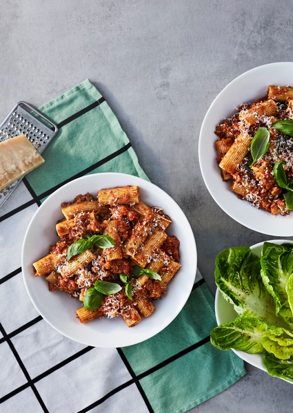 A bowl with pasta Bolognese made from HUVUDROLL plant balls minced, with fresh basil and grated parmesan, on a kitchen towel.