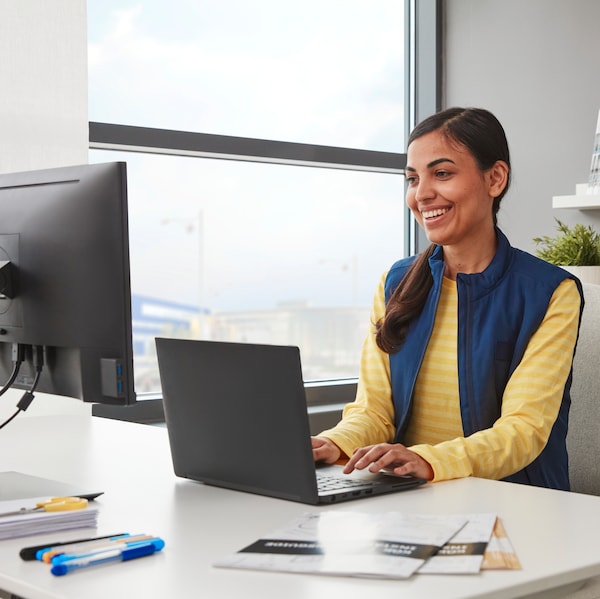 An IKEA co-worker dressed in yellow and blue sitting in front of a large white desk and computer smiling