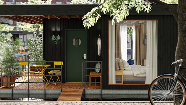 The deck of a small houseboat hotel floating on water in an urban context. One room with a bed can be seen through a window.