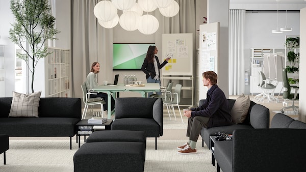 A young man sitting in a lounge area of several dark grey LILLEHEM sofa modules, TUNSTA tables and a meeting area behind it.