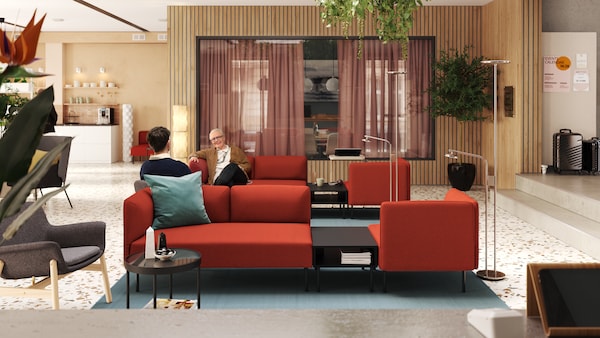 Two people relaxing in a modern, light-filled lobby and meeting room that has two red LILLEHEM modular sofas in the centre.