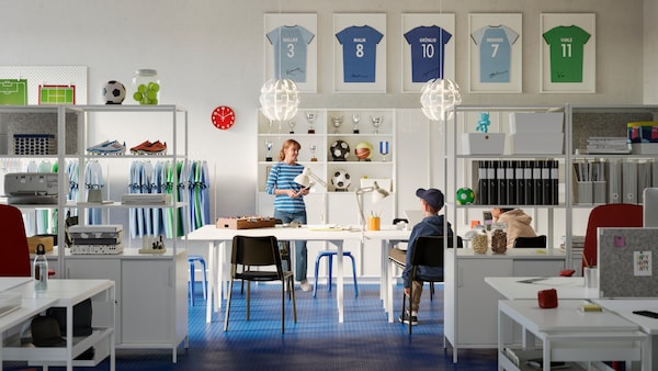 A large office with white TROTTEN tables and open storage shelves, black chairs and blue stools. Two people talking.