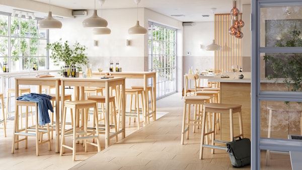 A bright and airy multi-zone bar with birch RÖNNINGE bar stools and tables by a window and seats at a bar in the centre.