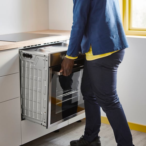 A man wearing a blue jacket and a yellow long sleeve tshirt installing an oven into a kitchen base cabinet.