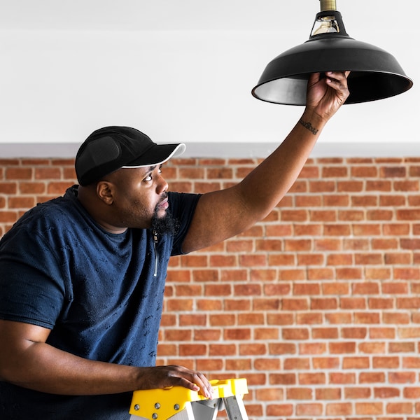 A man wearing a dark blue t-shirt is standing on a yellow ladder and screwing a light bulb in a black ceiling lamp shade.