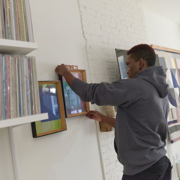 A man wearing a green long sleeve t-shirt and jeans mounting picture frames above a white bed.