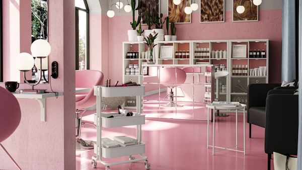 An airy pink hair salon with white KALLAX shelving behind a mirrored reception desk. A RÅSKOG trolley is by a workstation.