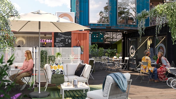 The sun is shining on an outdoor lounging area in a container village. People are socialising around grey SUNDSÖ tables.