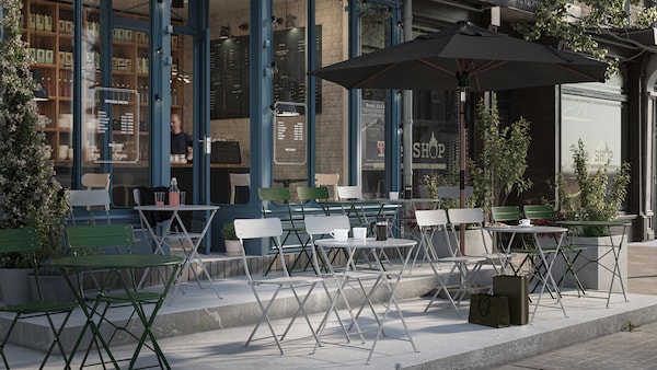 The terrace of a café with grey and green SUNDSÖ chairs and tables in lines facing outwards. An open parasol is on one side.