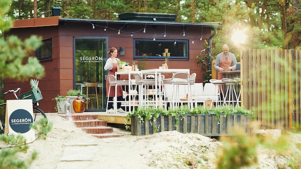 Two people are setting up an outdoor pop-up restaurant in the forest consisting of SEGERÖN outdoor furniture.