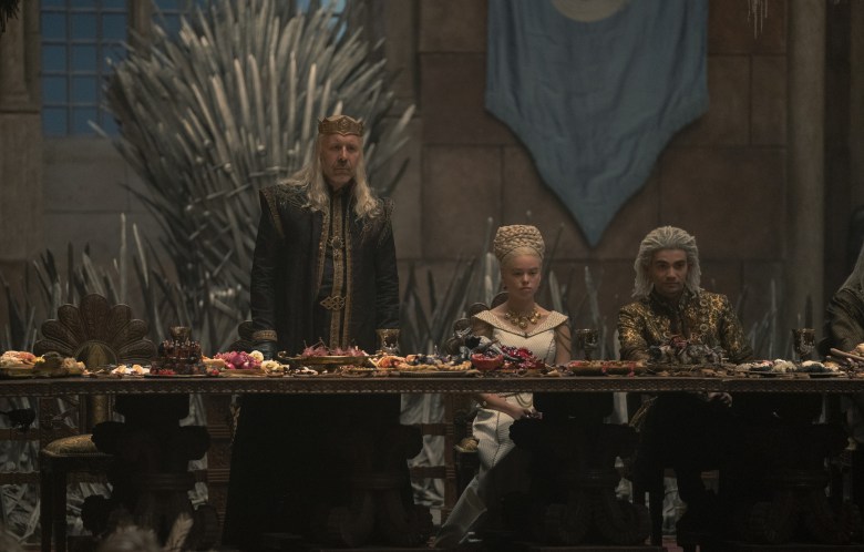 Three people at a long table in front of an iron throne; a man with long blonde hair, wearing a crown; his daughter in a white wedding dress with an elaborate blonde updo; the groom wearing gold and black; still from "House of the Dragon."