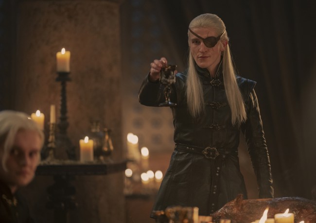 A young man with long, white-blond hair wearing medieval formalwear and an eye patch, raising a glass in a banquet hall full of candles; still from "House of the Dragon."