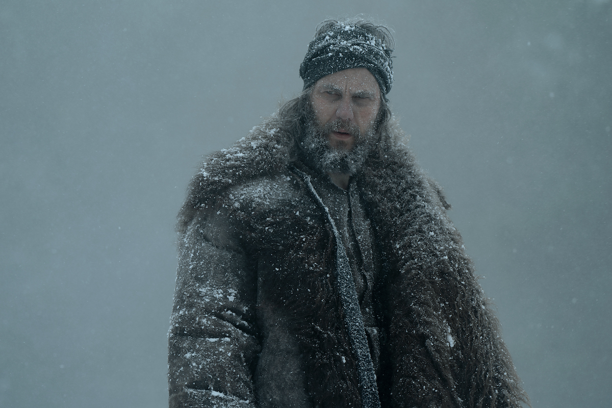 AMERICAN PRIMEVAL stars Taylor Kitsch as Isaac, shown here standing in the snow with a big furry coat and a big fuzzy beard