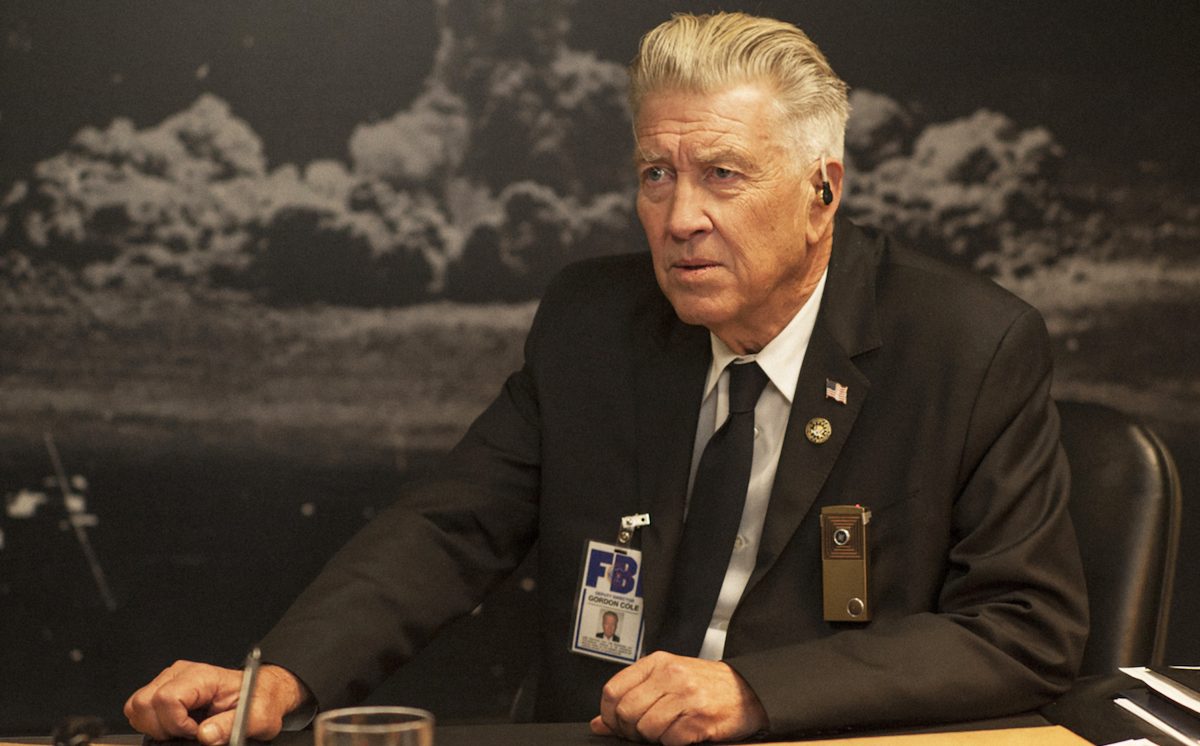 David Lynch in 'Twin Peaks: The Return,' sitting behind a desk, in front of a black-and-white photo of the atom bomb explosion, as Gordon Cole, FBI Agent