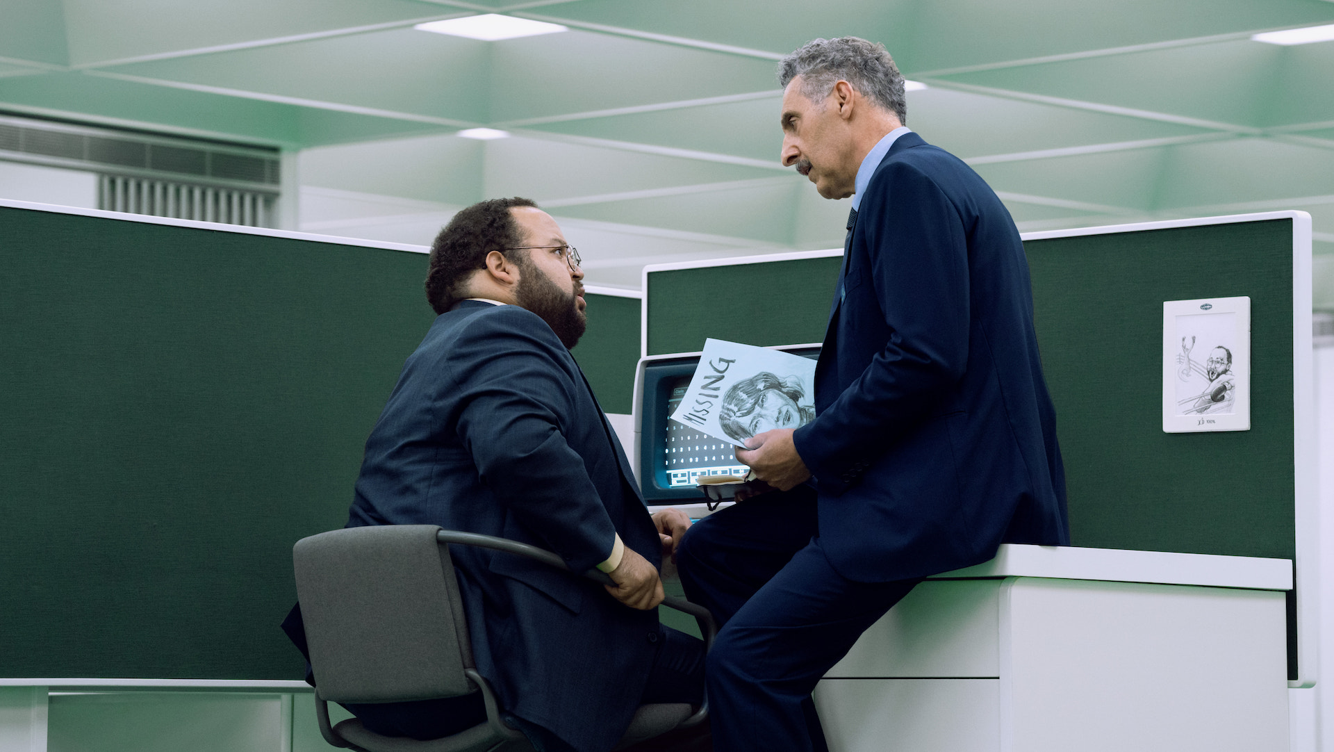 A man sitting at a desk and another sitting on it; Zach Cherry and John Turturro in 'Severance' Season 2