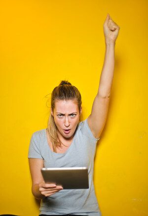 A photo of a woman holding an ipad and raising her hand in joy, as though she has just received good news.